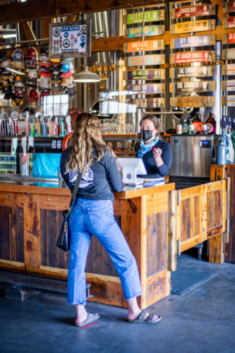 A woman stands in line behind a plexi-glass shield while a Draught Works server takes her order at the bar.